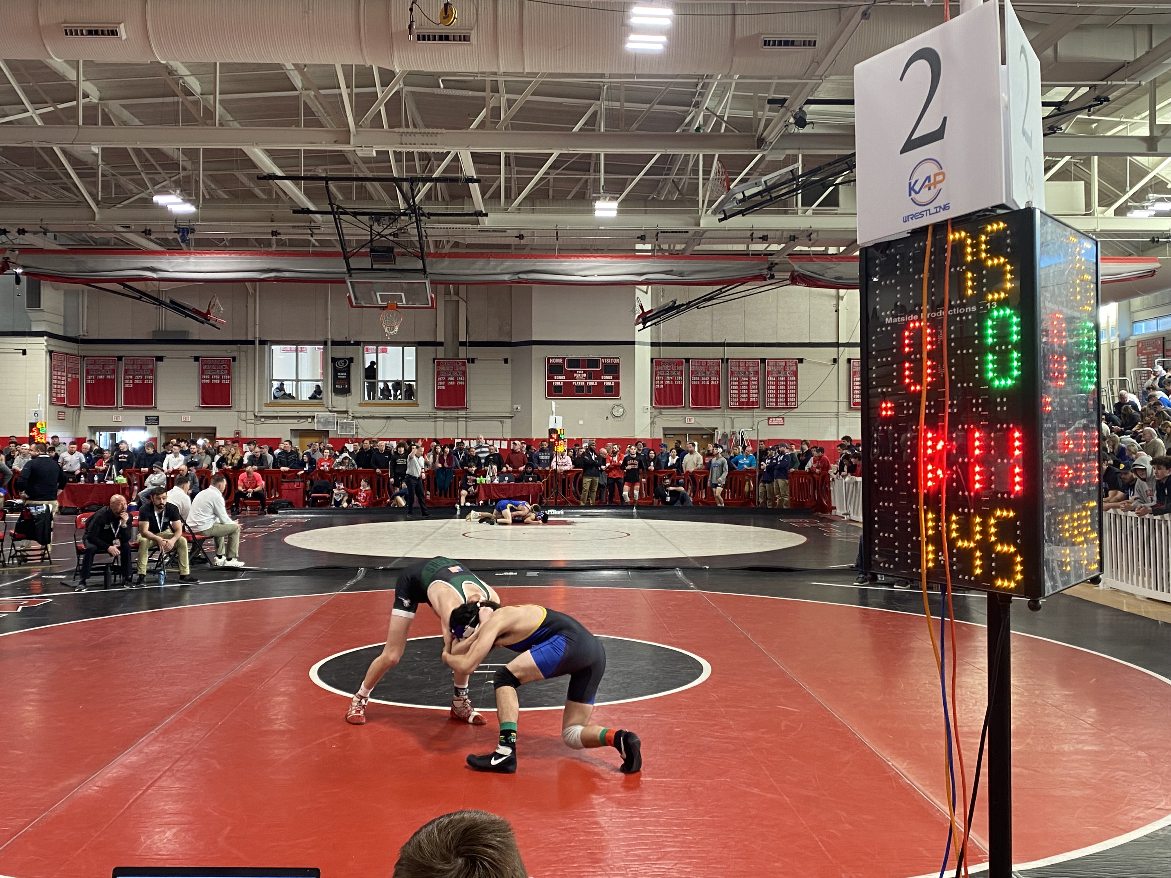 Two wrestlers compete on a mat in a gymnasium filled with spectators. The wrestler in green and black is grappling with the wrestler in blue and yellow. A large digital scoreboard shows the score and timer, with '2' prominently displayed at the top. Coaches and officials are seated around the mat, and the gym is decorated with red and black banners.