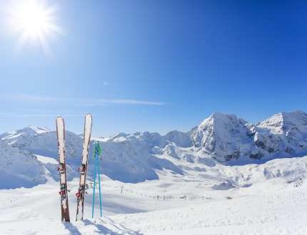 a pair of skis and ski poles dug into the snow with huge mountains and the bright sun behind