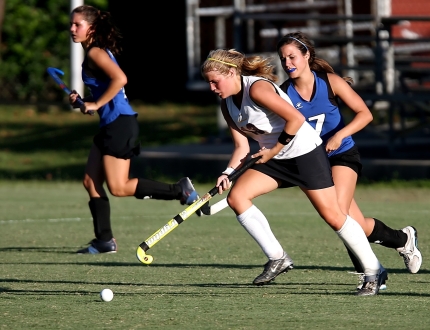 three players going after the ball in a field hockey game